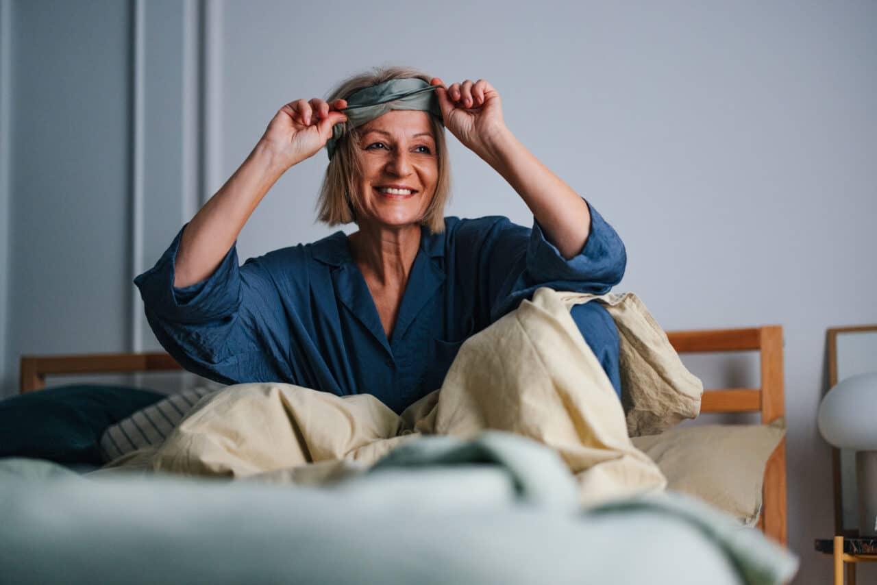 Woman in a comfortable bed, smiling and removing her eye mask after a restful night's sleep.