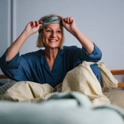 Woman in a comfortable bed, smiling and removing her eye mask after a restful night's sleep.